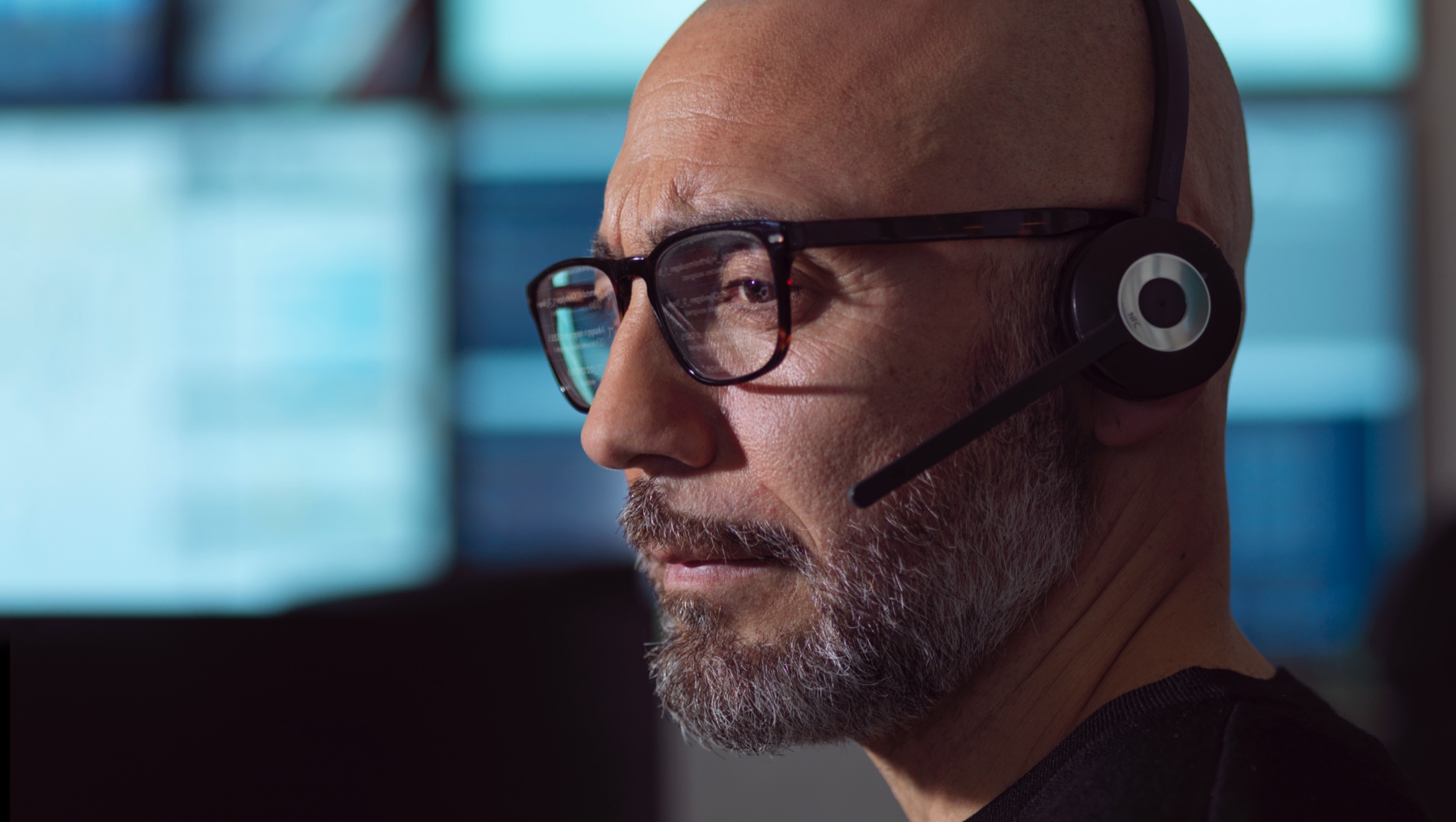 Primer plano de hombre con lentes y auriculares con micrófono mirando el monitor de una computadora.
